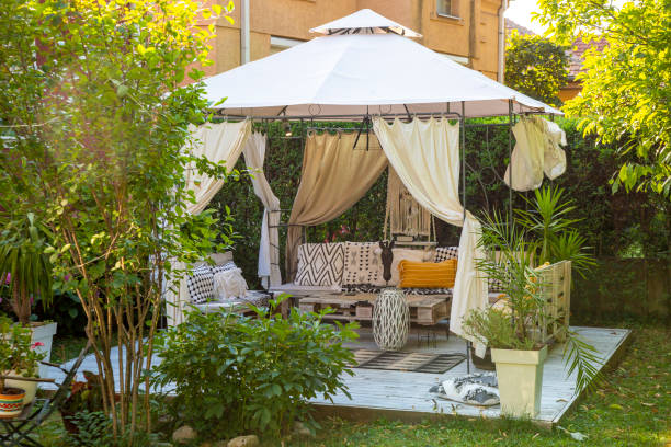 Beautiful cozy gazebo in the backyard surrounded with trees, flowers, lawn and fence at summer morning.