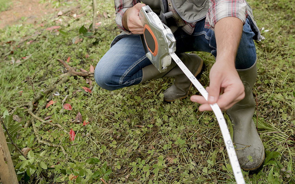Person in boots using a measuring tape to mark garden dimensions on grassy ground