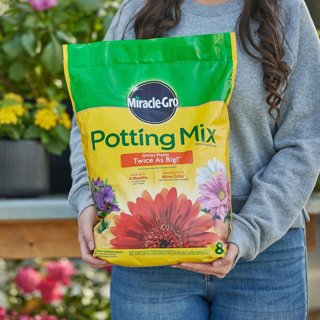 A woman carrying a bag of miracle grow potting mix in the foreground of a rack of potted plants.