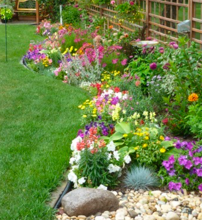 A vibrant garden border filled with colorful flowers such as petunias, snapdragons, and marigolds running along a green lawn beside a wooden trellis fence