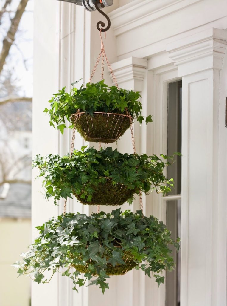 A three-tier hanging basket filled with cascading ivy and green foliage, suspended from a decorative hook near a white porch exterior.