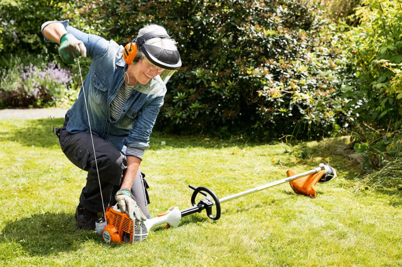 A older man cranking a gasoline operated edge trimmer with ear muffs protecting his ears, Gloves protecting his hands, long clothing protecting his body, and a face shield and goggles protecting his face and eyes.