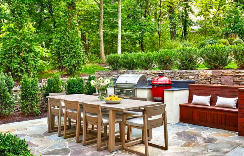 Backyard outdoor kitchen with built-in grill, kamado smoker, and dining set on stone patio.